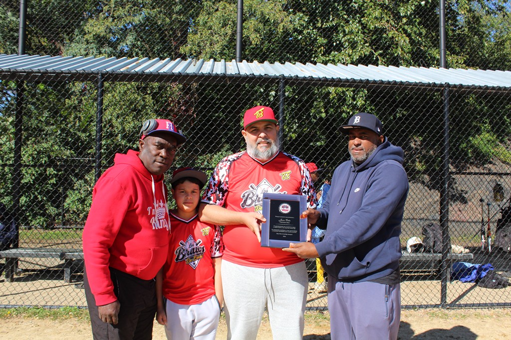 Juan Ramón Ortiz, acompañado de su hijo Christopher, recibe la placa de reconocimiento de manos de Ernesto de la Cruz, director de la Liga Los Bravos, y del coach José Maldonado, en agradecimiento por sus valiosos aportes al desarrollo del béisbol infantil y juvenil en El Bronx.